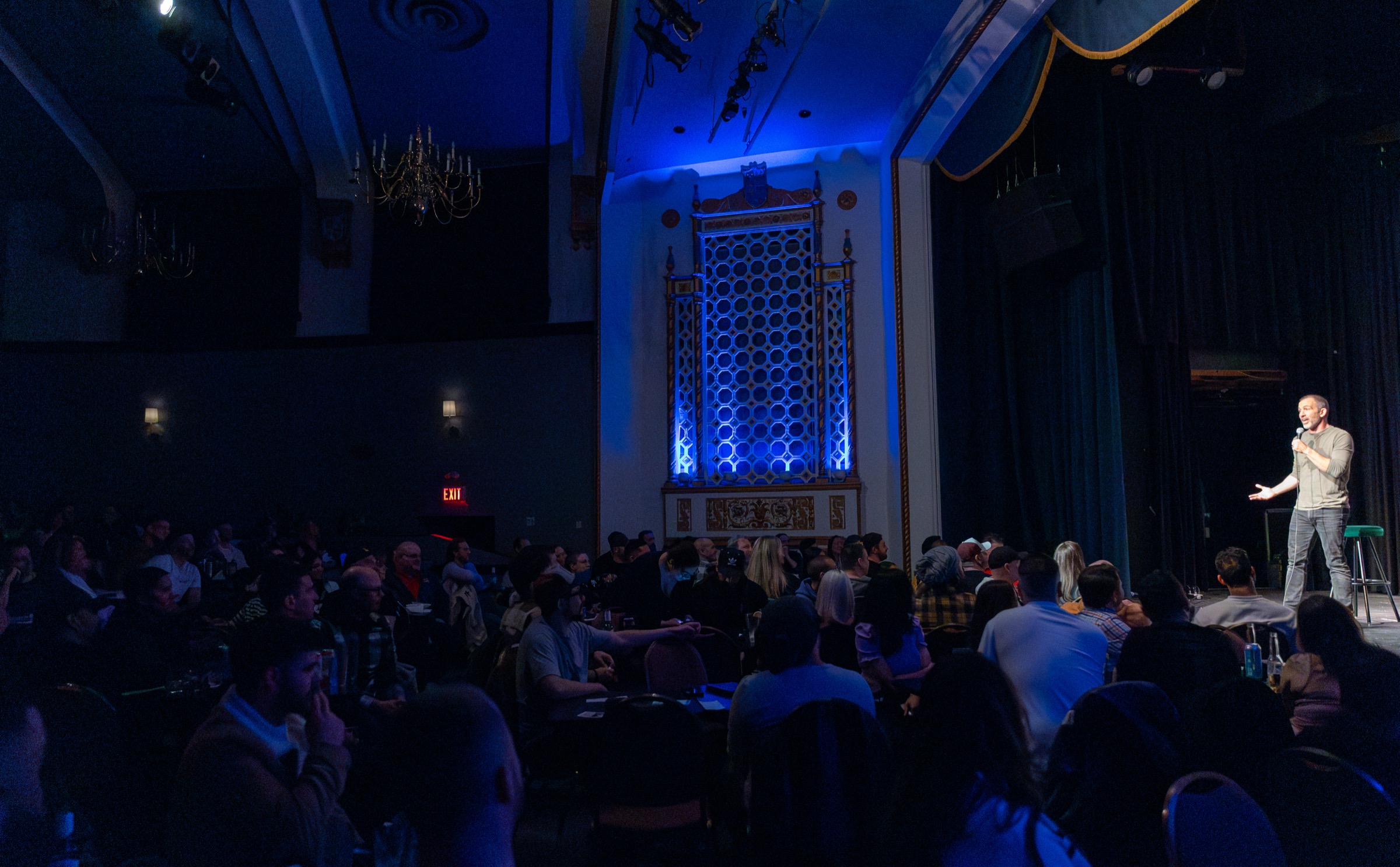 A comedian on stage at House of Comedy Vancouver in front of a full room, the venue's blue honeycomb wall glowing behind the crowd